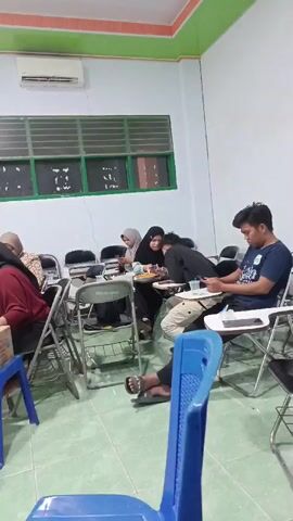 A video shows a group of people in a classroom setting. Several individuals are seated at tables, some of whom are wearing hijabs. One person in the foreground is focused on a mobile device. Another person is actively writing or taking notes. The room appears to be well-lit, with an air conditioning unit visible on the wall. The overall atmosphere suggests an educational or learning environment.
