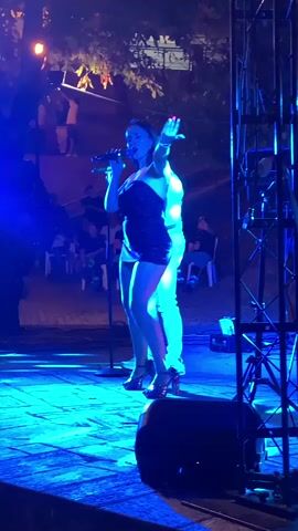 A woman is performing on a stage in front of an audience. She is wearing a short black dress and high heels and is holding a microphone. There is a man behind her. The stage is lit with blue and yellow lights. There are trees and lights in the background, and an audience is seated in chairs.