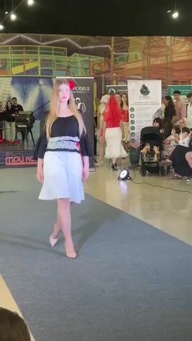 A young woman with long blonde hair and a red flower in her hair walks down a blue runway. She is wearing a black off-the-shoulder top with flared sleeves and white trim on the neckline and midsection, and white shorts. She is wearing nude high heels. In the background, there are several people watching and some banners. The environment appears to be an indoor event space with some colorful structures in the distance.