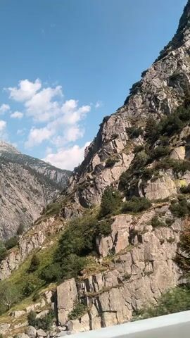 The video shows a mountainous landscape with a steep, rocky slope on the right and a lower, grassy slope on the left. There are trees and bushes growing on the slopes. In the distance, there are more mountains. The sky is blue with some white clouds. A body of water, likely a river, is visible at the bottom right of the frame. The camera appears to be moving, possibly from a vehicle, as indicated by the slight blur and the perspective shift. The speech in the video is in Russian, expressing fear and awe at the scenery and the road.