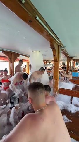 This video captures a vibrant foam party taking place on the interior deck of a boat, likely a tourist vessel, in Menderes, Turkey. A large group of young adults, both men and women, are seen enjoying themselves amidst a copious amount of white, bubbly foam that fills the entire floor space of the boat.

Most of the participants are wearing swimwear or light clothing, with many shirtless men and women in bikinis or swimsuits. They are actively engaged in the party, dancing, laughing, and interacting with each other as the foam is continuously generated and sprayed into the area from a large white inflatable or plastic structure positioned centrally. The atmosphere appears energetic and joyful.

The boat's interior features wooden beams and paneling, contributing to a rustic yet lively setting. Through the open windows or side railings, glimpses of bright blue water and a clear sky can be seen, indicating it is daytime and likely good weather. Some wooden benches or tables are visible along the sides, also partially covered in foam, with a few people sitting or leaning on them. The overall impression is one of a fun and festive summer activity at sea.