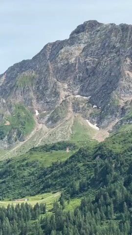 This is a vertical video of a mountain landscape. The foreground is blurred green foliage, likely trees. The middle ground shows a steep, grassy mountainside dotted with trees, and a patch of lighter green, possibly a meadow or field, with a few small structures visible. The background is a large, rocky mountain with visible patches of snow and scree slopes. The sky is a pale blue with some lighter clouds. The video appears to be captured from a moving vehicle or drone, indicated by the motion blur.