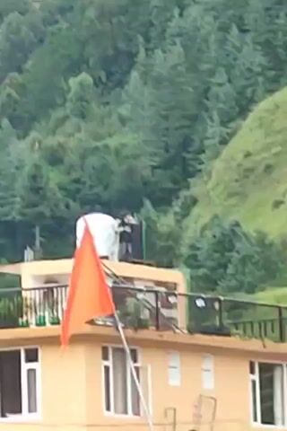 A brief video shows a rooftop scene with a water tank and an orange flag. Two individuals are standing near the water tank, engaged in an interaction. The background consists of a lush, green, tree-covered mountainside with some buildings visible in the distance. The weather appears overcast.