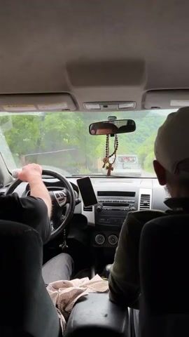 This media file captures the interior view of a car driving through a mountainous, possibly rural, area in Pasanauri, Georgia. The focus is on the road ahead and the two people inside. The driver, whose arm is visible gripping the steering wheel, is wearing a dark t-shirt. Beside them, a passenger wearing a white baseball cap and a dark green jacket is visible from behind.

The car's dashboard is in the foreground, featuring a phone mounted on a holder and a rosary hanging from the rearview mirror. The rosary is a notable detail, suggesting a religious element.

Through the windshield, a road is visible winding through a landscape characterized by lush green trees and hills. Buildings line the roadside, hinting at a small town or village. The weather appears to be overcast or rainy, with a slight blur on the windshield suggesting moisture. The time of day seems to be daytime, given the ambient light.

Visible text includes a warning sign on the sun visor and a sign on the roadside that reads "PASANAURI RESTAURANT." The overall atmosphere appears to be one of travel, perhaps a scenic drive through the Georgian countryside. There are no clear indications of strong emotions, but the activity is clearly focused on the journey itself.