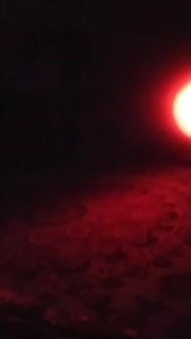 The scene is a dimly lit room, illuminated primarily by a red light source positioned towards the right side of the frame. The floor appears to be covered with a patterned carpet, with the red light casting a glow over the immediate area. There is significant darkness on the left side of the frame. The only distinct object is the light itself. There are no people visible.