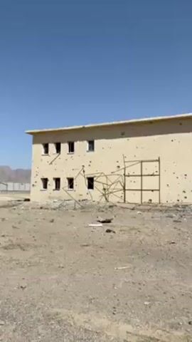 The video shows a battered two-story building. It is painted a light tan color and is riddled with bullet holes. Several windows are missing or broken. There is a rudimentary metal scaffolding structure propped against one wall. Debris is scattered on the ground in front of the building. The area appears dry and dusty. The sky is clear and blue. A man wearing a tan robe and red cap walks towards the building. Two other men are visible in the background near the building's entrance.