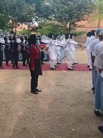 The video shows a group of young people in uniform standing at attention outdoors. They appear to be participating in a military-style drill or parade. In the foreground, a person in a red uniform stands facing the camera. Behind them, a larger group of individuals in white uniforms march in formation. A person in a blue patterned shirt and black pants walks across the frame, interacting with some of the marchers. The environment is an outdoor space with trees and buildings in the background. The ground is covered in dirt and gravel.