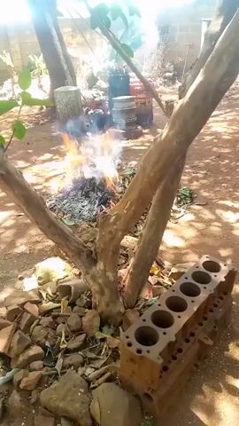 The video depicts an outdoor scene. A small fire burns on the ground, with a tree appearing to grow directly through the center of the flames. Nearby, resting on the ground, is a rusty engine block with six cylindrical openings. The ground is a mixture of dirt, small rocks, and fallen leaves. The light is bright, and the overall setting suggests a rural or outdoor workshop environment.