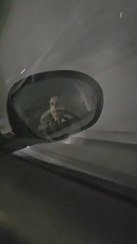 The video shows a driver as reflected in the side mirror of a car. The driver, a man, is facing forward and appears to be concentrating on the road. The car seems to be moving, as suggested by the blurred background. The ambient lighting is dim, possibly indicating that it's nighttime or the car is in a tunnel. The mirror's casing is dark, and there are light streaks on the mirror, perhaps from oncoming headlights or streetlights. The overall setting suggests the driver is alone, focusing on navigating the vehicle at night.