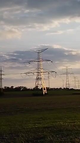 A static shot shows a field with a row of tall, metal electricity pylons extending into the distance. The sky is partly cloudy with a warm glow, suggesting either sunrise or sunset. The foreground is a grassy field. The pylons are evenly spaced and appear to be carrying high-voltage power lines. The scene is tranquil and devoid of human activity.