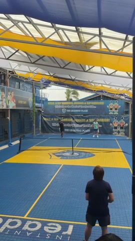 The video shows a pickleball game being played indoors under a blue and yellow canopy. Two people are on one side of the net, and one person is on the other. The person on the far side is wearing a green shirt and white shorts. The two people on the near side are wearing black shirts and shorts. The court is blue with yellow lines. There is a banner in the background with text and cartoon characters. The person on the right side of the court is in the process of hitting the ball. The person on the left side of the court is watching the play. The person closest to the camera is also watching the game. The court appears to be outdoors, but is covered by a tarp or awning.