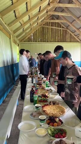 A group of men and one woman are gathered around a long table laden with food and drinks. They appear to be in an outdoor covered area with a wooden roof and corrugated metal walls. The table is covered with a white tablecloth and features various dishes, including flatbreads, fruits, meats, and bottles of beverages. The individuals are mostly looking down at the table, some are reaching for food, and one person is holding up a phone, possibly to take a picture or record. The atmosphere seems informal, possibly a picnic or a casual gathering.