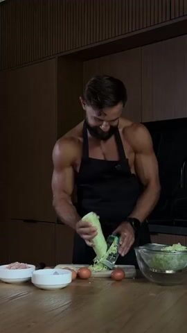 A man is in a modern kitchen, grating a zucchini into a bowl. He is wearing a black apron and is shirtless, showing off his muscular physique. There are other ingredients on the wooden countertop, including two bowls of what appears to be ground meat and chopped onions, as well as three eggs. A large glass bowl filled with grated zucchini is to the right of the man. The kitchen has dark wood cabinets and a black countertop. The lighting is dim, creating a dramatic effect. The man appears focused on his task, with a slight smile on his face. The video seems to be a close-up shot of the man's upper body and the ingredients he is working with.