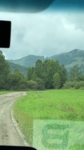 The video shows a dirt road curving through a green field. Tall trees are visible on the right side of the road, and rolling hills covered in vegetation are in the background. The sky is overcast and appears to be cloudy. A black rectangular object is affixed to the upper left corner of the windshield, obscuring part of the view. The footage seems to be captured from inside a vehicle.