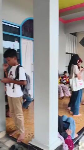 The video shows a group of people, mostly young, gathered in an outdoor covered area. Two young women in headscarves are standing and talking, one on the left wearing a tan shirt and blue jeans, the other on the right wearing a white headscarf and light blue pants. A young man with a grey backpack walks away from the camera towards an open doorway. Several other young people are sitting on the floor and on a bench in the background. A motorcycle is parked on the right side of the frame. Numerous pairs of shoes are scattered on the ground in front of the covered area. The environment appears to be a residential area.