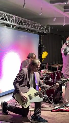 In this video, a band is performing on a stage. A male bass guitarist, wearing a brown suit and glasses, kneels on the floor as he plays a white bass guitar. Behind him, a drummer plays a red drum set. Another person, possibly a guitarist, stands to the right in a light pink suit. A large screen behind them displays a colorful, abstract pattern with pink and purple hues. The setting appears to be indoors, possibly a venue or event space, with stage lighting and trussing visible overhead. The mood seems energetic, indicated by the musicians' focused expressions and dynamic poses. There is no visible text in the media.