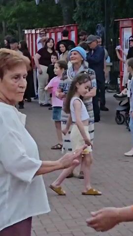 The video shows an outdoor scene where people are gathered. In the foreground, two women are visible, one in a white top and the other in an orange and purple patterned top. The woman in white appears to be looking at the woman in the patterned top. Further back, a woman wearing a blue headscarf and a patterned dress is dancing. She has her mouth open as if singing or exclaiming. Several other people, including children and adults, are standing or walking around in the background. They appear to be in a park or a public outdoor space, with trees and lamp posts visible. There are also red structures that resemble booths or stalls in the background. The ground is paved with bricks.