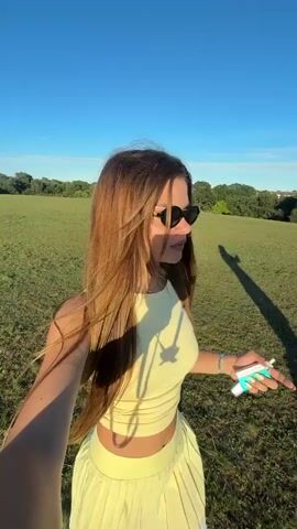 A young woman with long brown hair and sunglasses stands in a grassy field under a clear blue sky. She is wearing a yellow crop top and a pleated yellow skirt. She is holding a set of keys in her right hand. The sun is casting a long shadow of the woman on the grass.