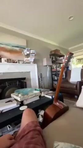 Here is a detailed description of the video:

The video starts from a low angle, showing the lower half of a person wearing red shorts sitting on a brown couch. The camera pans slightly upwards and to the right, revealing more of the living room. A brown acoustic guitar is leaning against the couch, positioned between the person and the viewer. A black coffee table is in front of the couch, covered with books, decorative items, and what looks like a white remote control.

Behind the coffee table, there is a white mantelpiece above a dark fireplace. The mantelpiece is decorated with numerous photographs and cards. To the right of the fireplace, a slender, silver floor lamp stands next to a small bookshelf filled with various items, including suitcases stacked on top.

Further to the right, an armchair with brown leather upholstery is visible. Natural light streams in through a window behind the armchair. The walls are painted in a neutral color, and the ceiling is white.

As the camera continues to pan upwards, it moves past the person and the ceiling is visible. The camera then abruptly changes direction, and a blurry image of a person's face is shown. The video then abruptly ends.