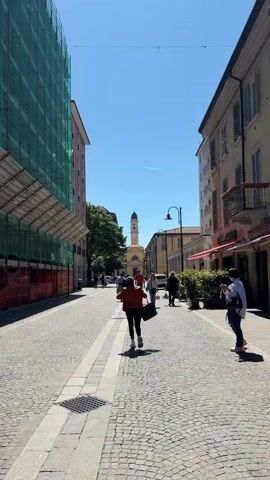 The video captures a street scene on a sunny day. The street is paved with cobblestones and has sidewalks on either side, lined with buildings. One building is under construction, covered in green scaffolding. Several people are walking along the street, mostly away from the camera, towards a church tower visible in the distance. There is also a person standing on the right side of the street, looking at their phone. The sky is clear blue.