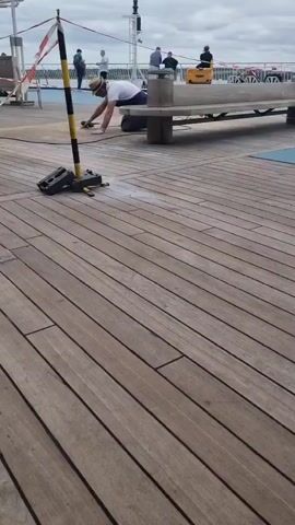 A man in a white t-shirt, light pants, and a hat kneels on a wooden deck, operating a handheld tool connected to a cable or hose, appearing to sand or clean the deck. A black and yellow striped pole with red and white caution tape is positioned to his left, with two black weights at its base. White dust or residue is visible on the deck where he is working. Several other people are standing and sitting on benches further in the background. The deck is composed of light brown wooden planks with dark seams. The sky is overcast.
