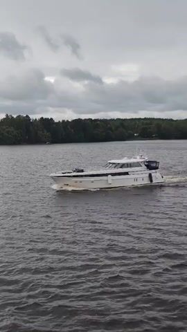 A white luxury yacht is cruising on choppy gray water. The yacht has a sleek design with multiple windows and a dark-colored enclosed bridge at the stern. A wake trails behind the boat. The background shows a lush green tree line along the shore under a cloudy, overcast sky. The camera appears to be on a moving platform, possibly another boat, capturing the yacht from the side.