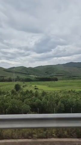 The video shows a view from a moving vehicle, looking out at a landscape. The foreground features a metal guardrail and some vegetation. Beyond that, there are green fields and trees, with rolling hills covered in vegetation in the background. The sky is overcast with grey clouds. There are also some tall power lines visible in the distance. The overall impression is a rural, natural landscape under a cloudy sky.