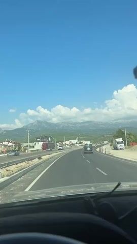 A first-person perspective video shows a view from the driver's seat of a car driving on a highway. The road is paved and has multiple lanes. There are other vehicles on the road, including cars and trucks. On the side of the road, there are construction barriers and some buildings. In the distance, there are mountains and a clear blue sky with white clouds. The dashboard of the car is partially visible at the bottom of the frame, showing speed and other indicators. The overall impression is a sunny day journey.