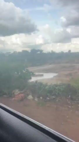 This media file captures a natural scene in Mangu Halle, Nigeria. The subject is a rocky riverbed with murky brown water flowing over smooth, light-colored stones. Tall, green grass grows along the foreground and to the right of the river. In the background, trees and distant structures suggest a rural or semi-rural setting. The sky is filled with dramatic, dark, and light gray clouds, indicating potentially unstable weather, possibly leading up to or following rain. The lighting suggests it is daytime, but the overcast conditions might make it appear darker than a clear day. The presence of what appear to be remnants of a wooden structure, possibly part of a bridge or railing, hints at human interaction with the natural environment, though no people are visible. The overall mood is serene yet somewhat dramatic due to the cloudy sky and the rugged landscape. There is no visible text in the media file.