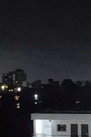 This is a low-light, shaky video of a city skyline at night. In the foreground, there is a white structure, possibly a building or a rooftop. In the background, there are several dark buildings, some with illuminated windows. The sky is dark and cloudy. There are no people or discernible actions visible in the video.