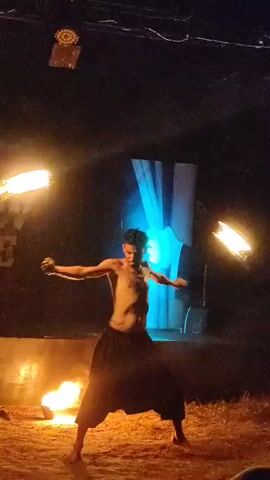 A shirtless man with dark, loose-fitting pants performs a fire-dancing routine outdoors at night. He is in the center of the frame, with flames visible on either side of him at ground level. He holds a flaming prop in each hand and is in the process of breathing fire, with a large plume of flame erupting from his mouth. The background is dark, with some stage lighting and a blurred banner visible. The ground is sandy or dusty.