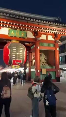 The video shows a nighttime scene at what appears to be a Japanese temple entrance. The large red gate with green and gold signage hangs a large paper lantern with black characters. Several people walk through the gate, some taking photos with their phones. The area is well-lit and crowded, with more temple structures visible in the background. Most people are wearing casual clothing. The overall atmosphere seems to be a mix of tourism and local activity.
