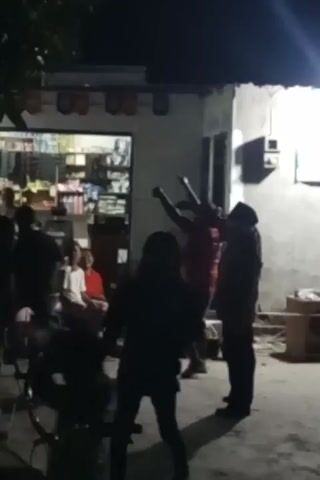A group of people are gathered outdoors at night. A man in a red shirt appears to be dancing or performing, raising his arms. Another person, silhouetted and wearing a hooded garment, stands opposite him, also with their arms raised. Several other people are seated or standing in the background, observing. Behind them is a building with shelves stocked with items visible through a window, suggesting it might be a shop. The ground is paved, and a bicycle is partially visible in the foreground. The lighting is dim, with artificial light sources illuminating the area.