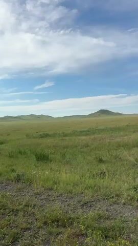 The video shows a landscape of rolling green hills under a bright blue sky with scattered white clouds. The camera is positioned inside a vehicle, looking out a side window, and a portion of the car's side mirror and the side of the road are visible in the foreground. The road appears to be unpaved or gravel. The vast, open grassland stretches to the horizon where the hills are located.