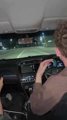 The video shows the interior of a car driving at night. In the driver's seat, a person's hands are visible on the steering wheel. To the right of the driver, a person's hands are on the center console, interacting with controls. In the passenger seat, a person wearing a light-colored hoodie with a black logo on the left chest is visible. This person is looking forward and then turns their head to the right, appearing to speak or react. The background outside the car shows streetlights and passing scenery, indicating movement on a road, likely a highway. The lighting is dim, consistent with nighttime driving.
