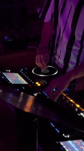 A DJ, wearing a white striped shirt, is actively working at a professional DJ mixer in a dimly lit environment. The mixer is equipped with a jog wheel that the DJ is manipulating with their right hand. The DJ's left hand is also visible and appears to be adjusting controls on the mixer. The mixer features multiple illuminated buttons and screens displaying audio information. The overall scene suggests a nightclub or performance setting.