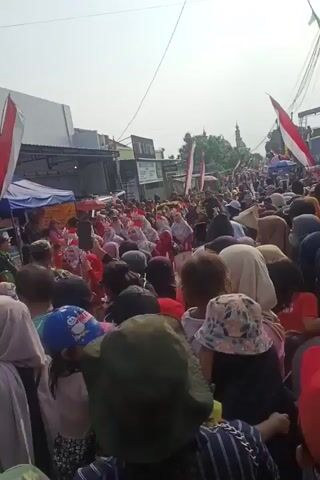 A crowded outdoor street scene. Many people are visible, mostly facing away from the camera. Several individuals are wearing red and white clothing, and some are holding small Indonesian flags. There are stalls or vendors set up along the side of the street. The sky is bright, suggesting daylight. The overall impression is of a public gathering or event.