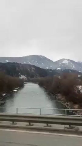The video shows a river running between bare trees with snow-covered mountains in the background, viewed from a bridge. The sky is overcast. The bridge's guardrail is visible in the foreground. The river appears calm with a greenish hue. The scene is stationary, taken from a fixed perspective.