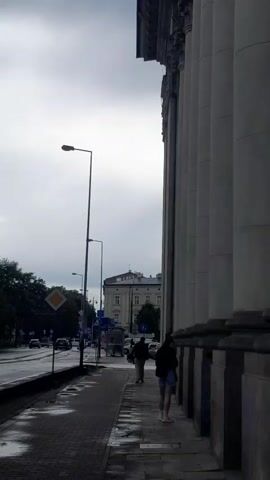 A vertical video displays a sidewalk next to a large building with columns. Several people are walking along the sidewalk. To the left, a street with traffic and a traffic light is visible. The sky is overcast and appears to be raining.