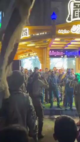 The video shows a group of people gathered in front of a building with Arabic writing and bright lights. Most of the people are standing and watching two men in traditional clothing dancing. The dancers are facing away from the camera, and their movements seem energetic. Many people in the crowd are holding up their phones, apparently recording the performance. The environment is lively, with music playing in the background. Some of the building's signage is in Arabic.