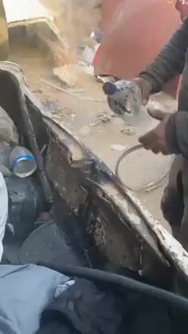 A man in a grey and black jacket stands in a junkyard next to what appears to be a vehicle frame. The frame is grey and black. There is debris scattered around the ground. The man pours water from a water bottle onto the frame. He then wipes the frame with his hand. The man smiles at the camera.