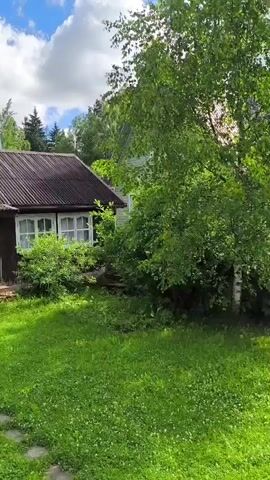 The video shows a rural landscape with a small wooden house. The house has a dark brown wooden exterior and a corrugated metal roof. There are white-framed windows and a small porch with steps leading up to the entrance. The house is surrounded by lush green grass and trees. In the background, there are more trees and a blue sky with white clouds. The video pans slowly to the left, revealing another wooden structure, possibly a shed or another small building, and more vegetation. The overall scene is peaceful and evokes a sense of rustic charm.