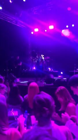 A concert is underway, illuminated by vibrant purple stage lights. In the center, a performer in a wide-brimmed hat and a colorful, patterned outfit with fringe accents is singing into a microphone. Behind them, a band is visible, including a drummer on a drum set. In the foreground, the backs of several audience members are visible, facing the stage. The atmosphere is energetic and celebratory.