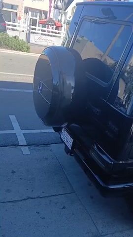 The video shows a dark-colored Mercedes-Benz G-Class SUV parked on a city street with shops and restaurants visible in the background. The camera moves around the vehicle, showing its rear, side, and front, highlighting details like the spare tire mounted on the back, the running boards, and the wheels. The SUV has tinted windows, and the license plate is visible. The surrounding environment includes street signs, other parked cars, and the building facades of businesses such as "Pure & Duda" and "RikBay". The weather appears to be sunny.