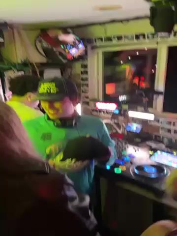 The video shows an indoor party with neon lights. A DJ in a teal shirt and backwards cap is playing music. A woman with long brown hair and sunglasses dances, raising her hands in the air. Other partygoers are visible in the background. The atmosphere appears lively and festive.