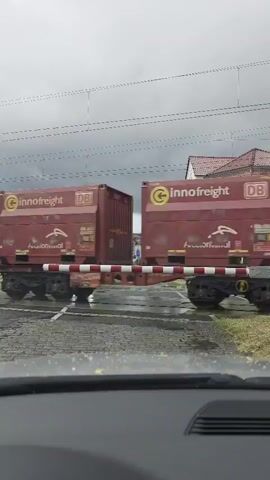 A train carrying two large cargo containers is passing through a railway crossing. The containers are red with white lettering that says "innoſfreight" and "DB." The train cars have white and red striped barriers across the crossing. The sky is overcast and it appears to be raining. The video is taken from inside a vehicle, as indicated by the dashboard in the foreground.