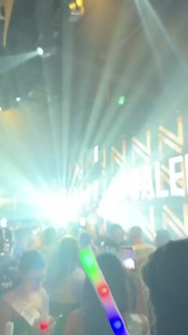 A female performer sings and dances on a stage in front of a crowd at a nightclub called Galeria. The crowd films her with their phones. She wears a glittery two-piece outfit. The backdrop is an LED screen displaying the word “GALERIA” and a geometric design that changes color. The club has a dark ceiling and a sound system. The lighting is colorful and changes as she sings. One member of the audience holds a light-up wand.