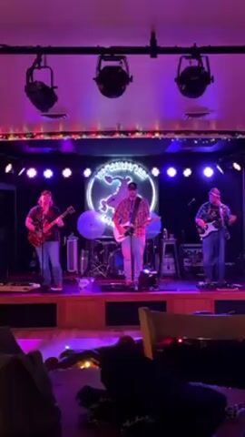 A band plays onstage in a bar. Three men stand on the stage, all holding guitars, and presumably singing. A neon sign behind them reads “THE COVERED WAGON.” The stage is illuminated by several spotlights and blue lights. Other equipment is visible behind the musicians, including a drum set. The foreground shows the corner of a table, covered with cloth. The room is dark, but well-lit.