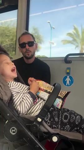 This video captures a man and a young child traveling inside a vehicle, likely a bus or train, in Khalde, Lebanon. The child, appearing to be between 2-4 years old, sits in a dark grey stroller, wearing a striped long-sleeved shirt and dark pants adorned with a space-themed pattern that includes the partial word "ADVENTUR." Several bags of snacks, prominently featuring one with the Hebrew text "במבה" (Bamba), rest on the stroller's front bar. The man, wearing a black long-sleeved shirt and sunglasses, sits behind the child. Through the large window, palm trees, bright sunlight, and buildings, including a white multi-story structure, indicate a clear, sunny daytime. A blue sign with a white pedestrian icon is visible on the interior wall of the vehicle. The child initially appears animated and vocal, then observes their surroundings with a thoughtful expression. The man maintains a neutral demeanor.
