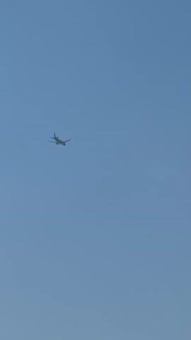 The video shows a small airplane flying in a clear blue sky. The airplane is a light-colored jet with its wings extended. It is viewed from below and slightly to the side, making it appear as a silhouette against the bright sky. There are no visible passengers or people on the ground. The setting appears to be outdoors, possibly during the daytime, given the brightness of the sky. There is a faint, dark circular object visible in the sky to the right of the airplane, its nature is unclear. The video does not contain any visible text. The activity is the flight of the airplane across the sky. No specific emotions are conveyed by the media.